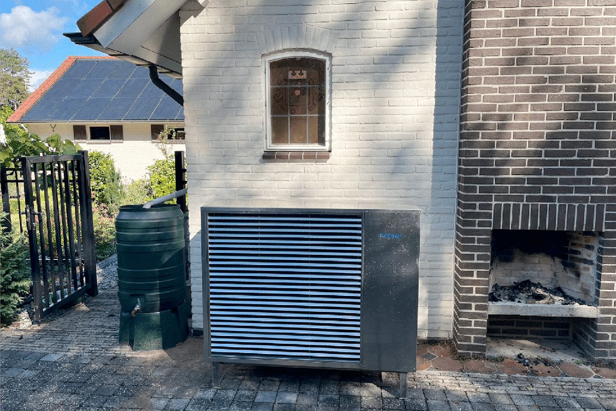 Buitenunit van een warmtepomp naast een regenton tegen een witte gevel